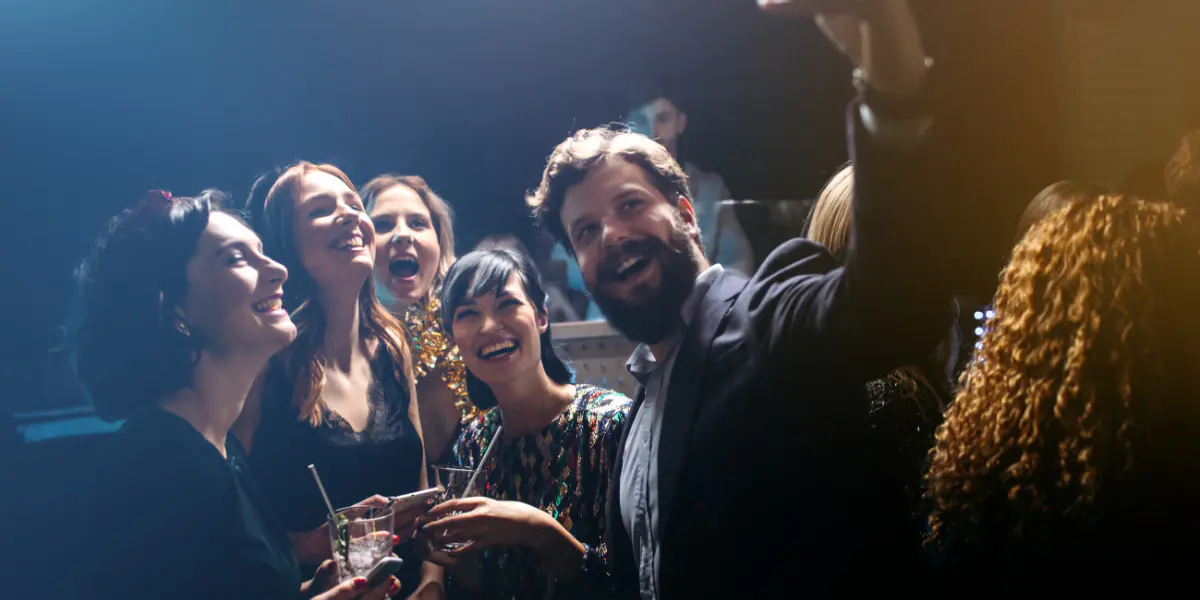 A group of smiling friends taking a selfie together at a crowded, dimly lit party or event.
